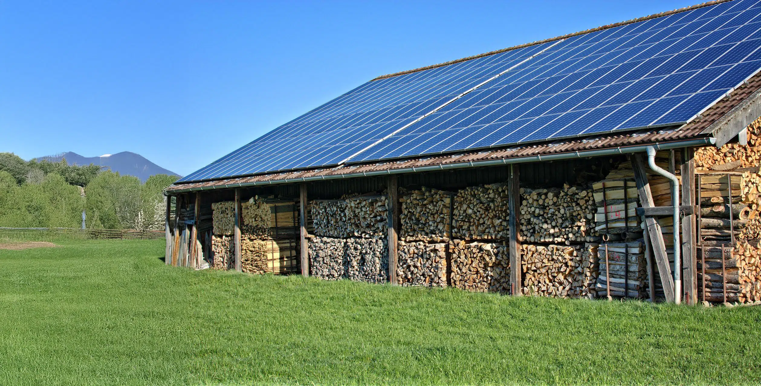 A wood store under a roof with solar panels, surrounded by green grass and with mountains in the background.